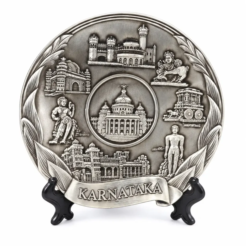 Silver-tone Karnataka souvenir plate with landmark and figure relief, leaf border and black stand on a white background