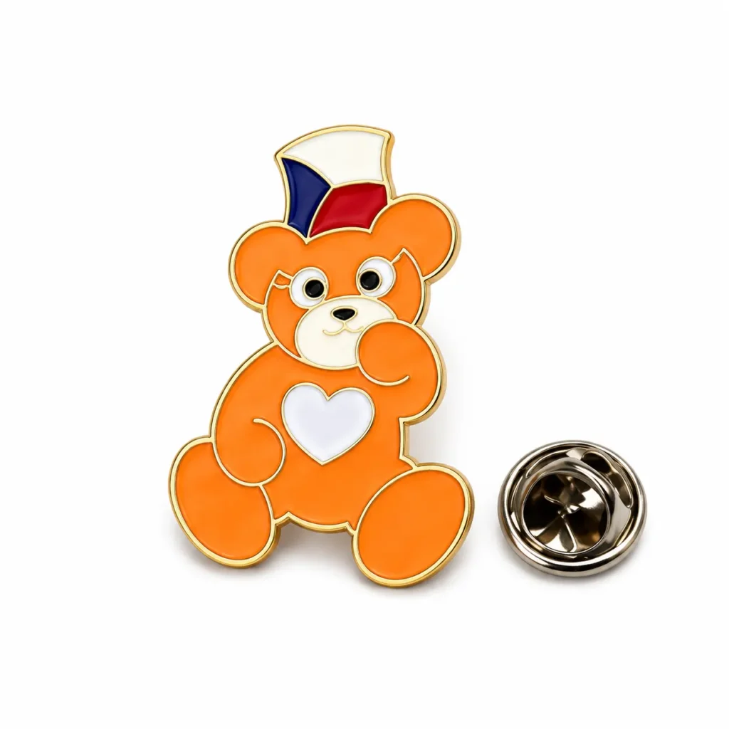 Orange teddy bear enamel pin with a blue, white and red hat, white heart and separate butterfly clasp